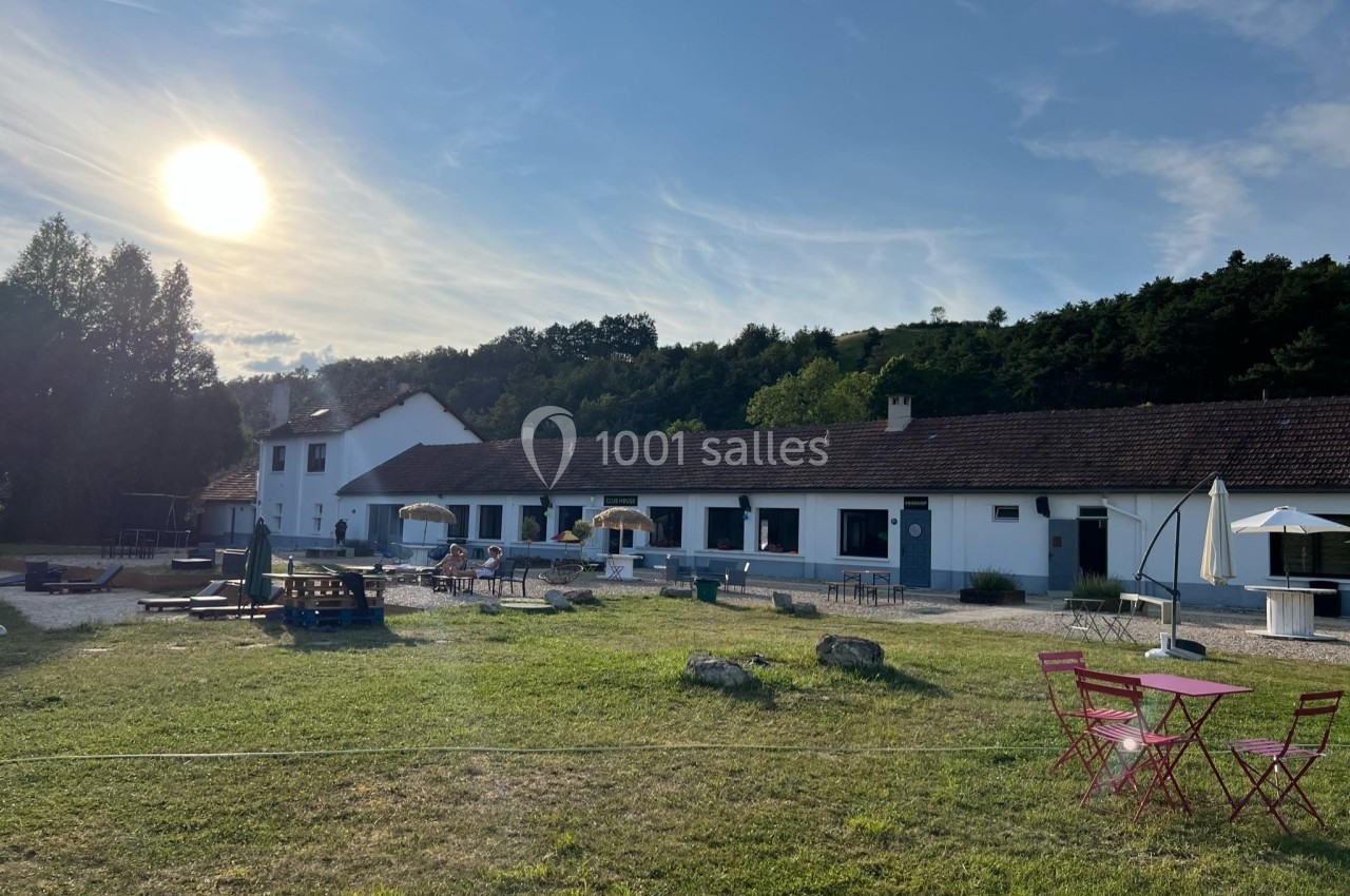 Bâtiment blanc de plain-pied entouré de verdure, avec tables extérieures et soleil bas dans le ciel.