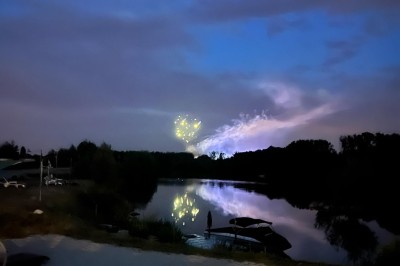 Vue d'une rivière bordée de végétation avec une maison au toit rouge et un ciel partiellement nuageux au coucher du soleil.
