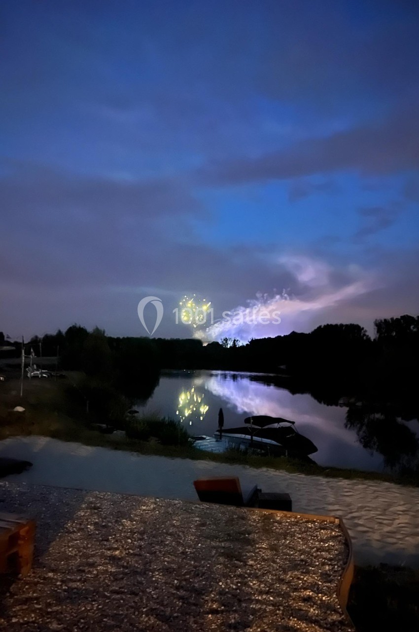 Ciel nocturne avec des feux d'artifice reflétés sur un lac calme, entouré de végétation et d'un rivage éclairé.