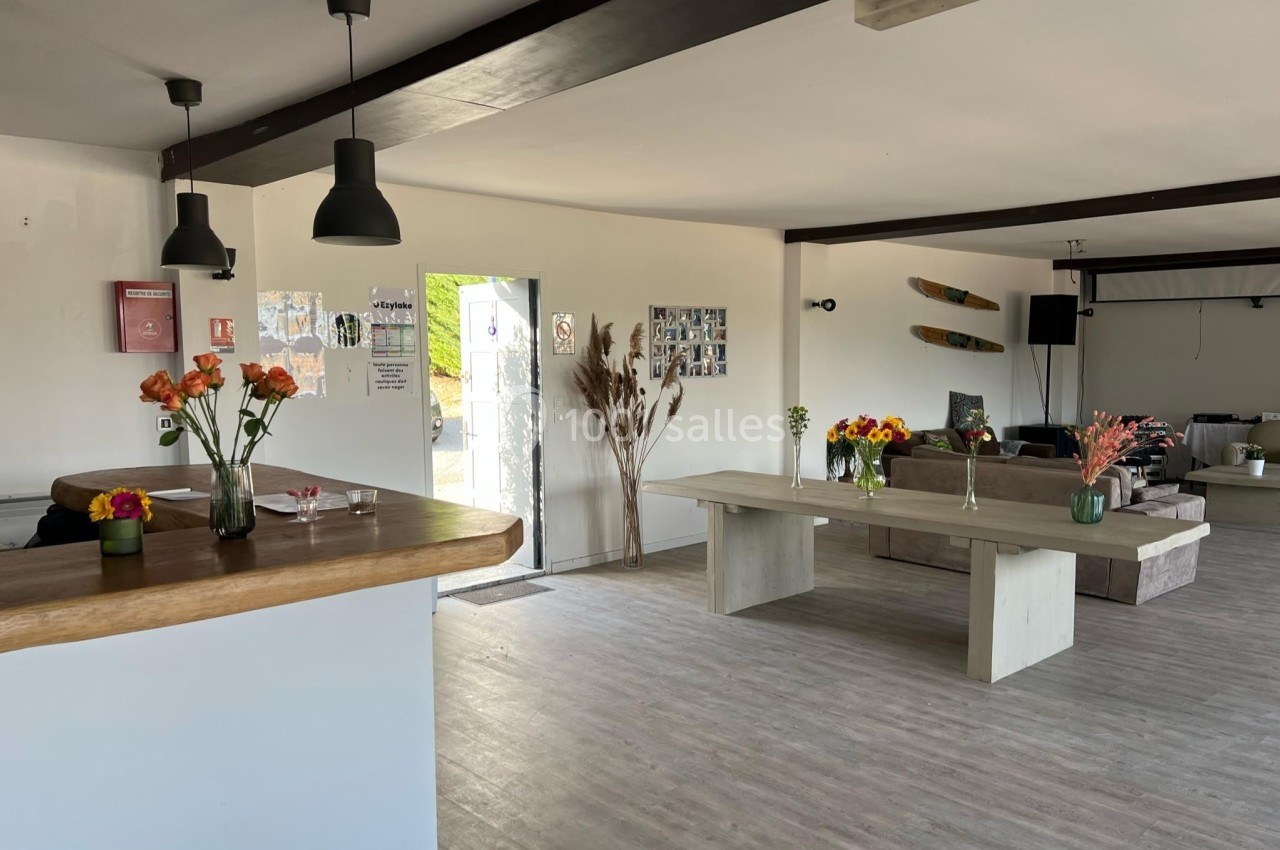 Salle lumineuse avec tables en bois, bouquets de fleurs, canapés et décoration murale, vue sur une porte ouverte.