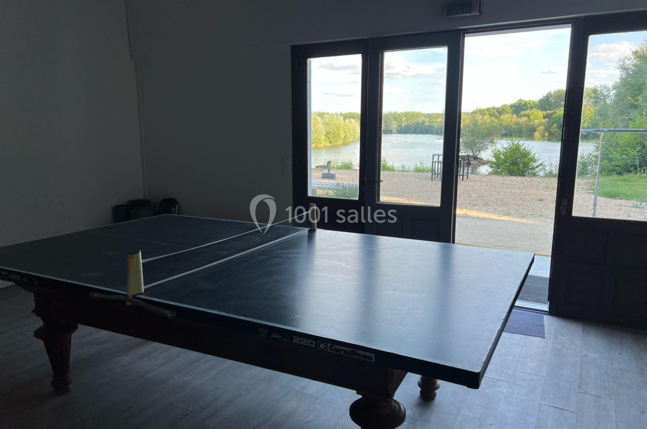 Table de ping-pong dans une pièce sombre avec vue sur un lac et des arbres à travers de grandes baies vitrées.
