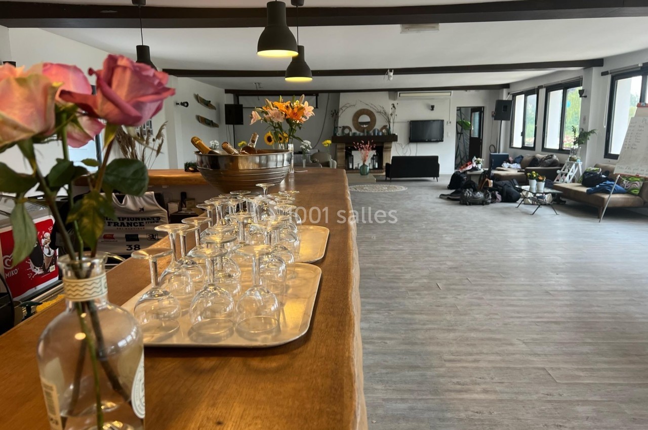 Salle lumineuse avec un comptoir en bois, des verres alignés, des fleurs en vase et un espace détente en arrière-plan.