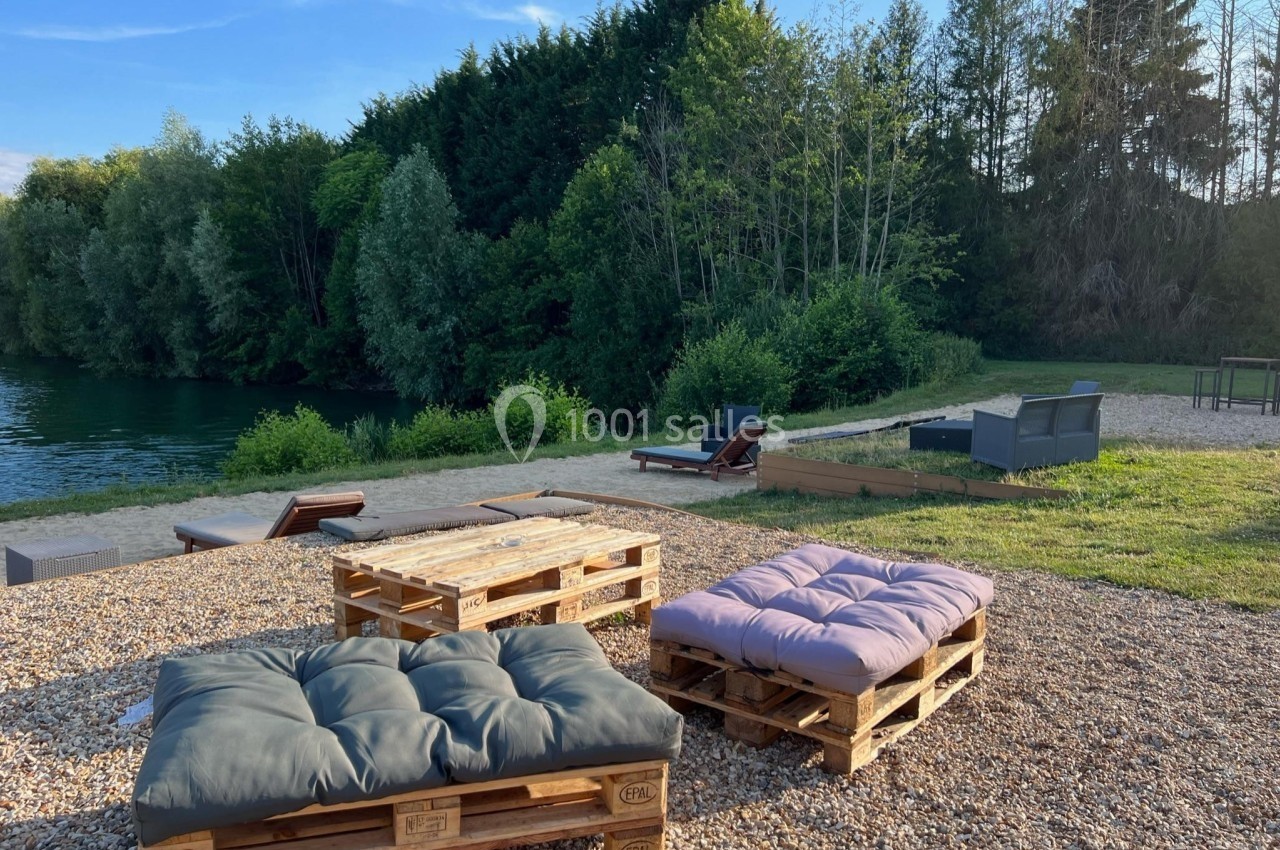 Mobilier en palettes avec coussins installé sur un sol gravillonné, près d'un plan d'eau entouré de végétation.