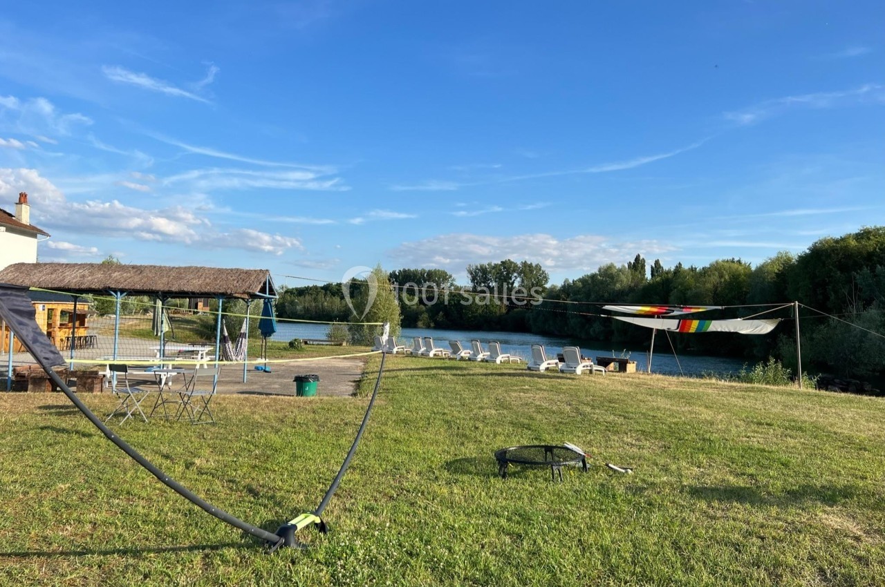 Espace extérieur avec pelouse, chaises longues, terrain de volley et vue sur une rivière bordée d'arbres.