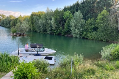 Vue d'une rivière bordée de végétation avec une maison au toit rouge et un ciel partiellement nuageux au coucher du soleil.