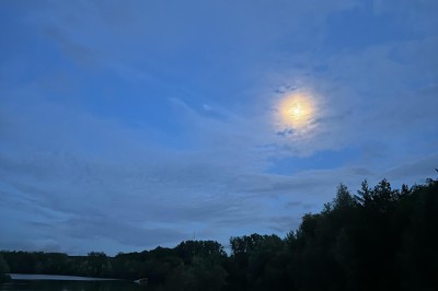 Vue d'une rivière bordée de végétation avec une maison au toit rouge et un ciel partiellement nuageux au coucher du soleil.