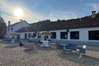 Vue d'une rivière bordée de végétation avec une maison au toit rouge et un ciel partiellement nuageux au coucher du soleil.