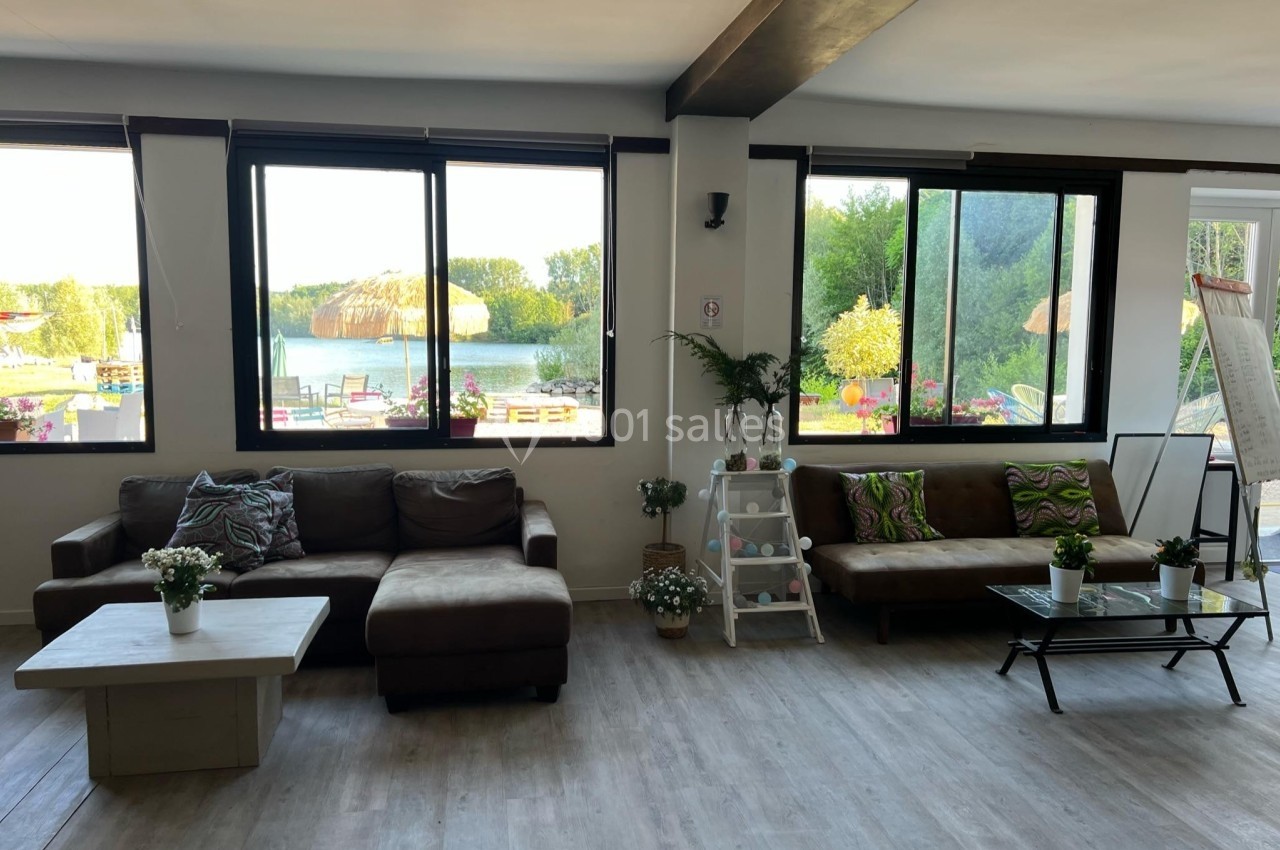 Salle lumineuse avec canapés, tables basses, plantes et grandes fenêtres donnant sur un paysage verdoyant.