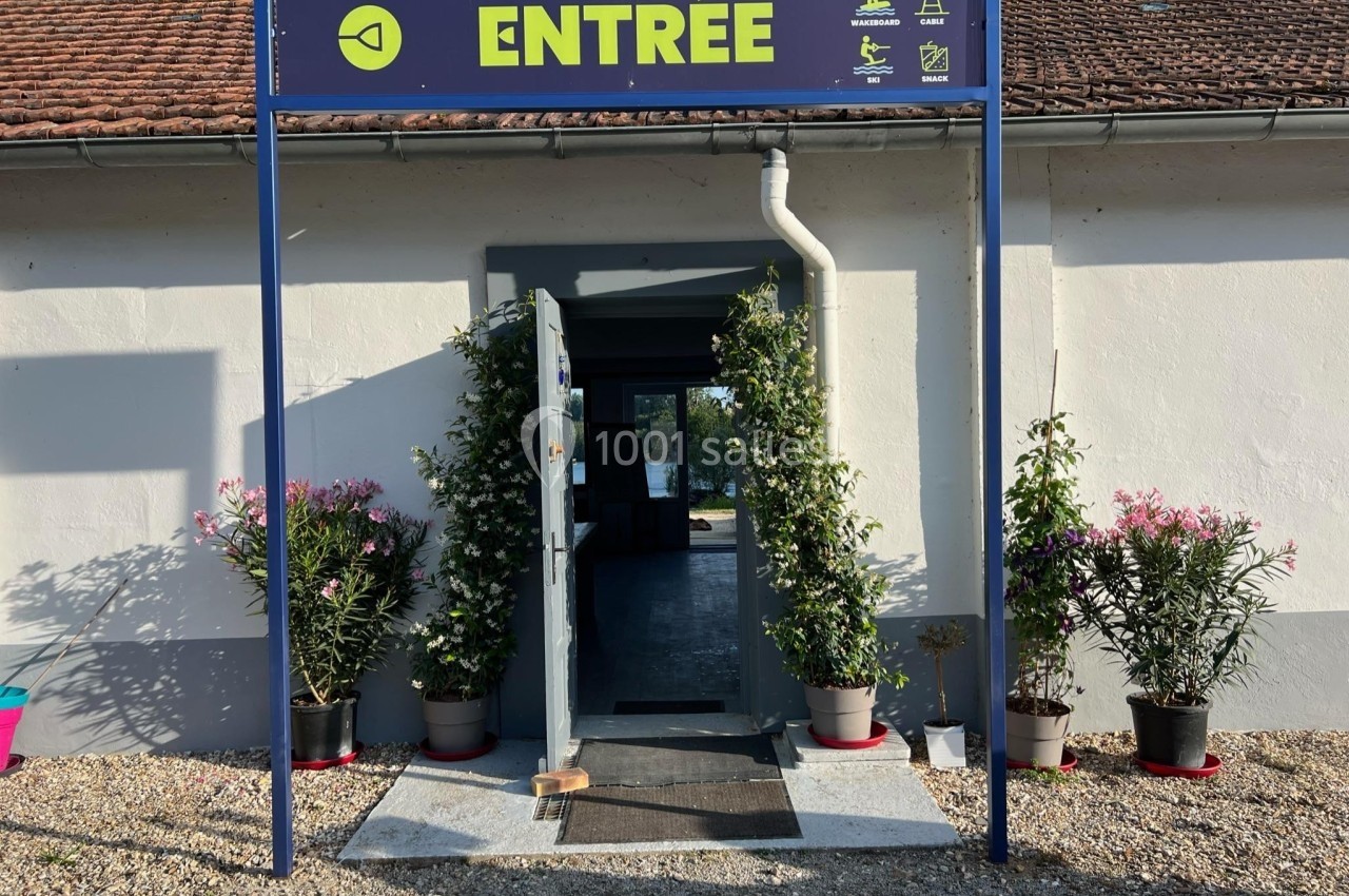Entrée d'un bâtiment avec porte ouverte, encadrée par des plantes en pot et un panneau indiquant ’Entrée’.
