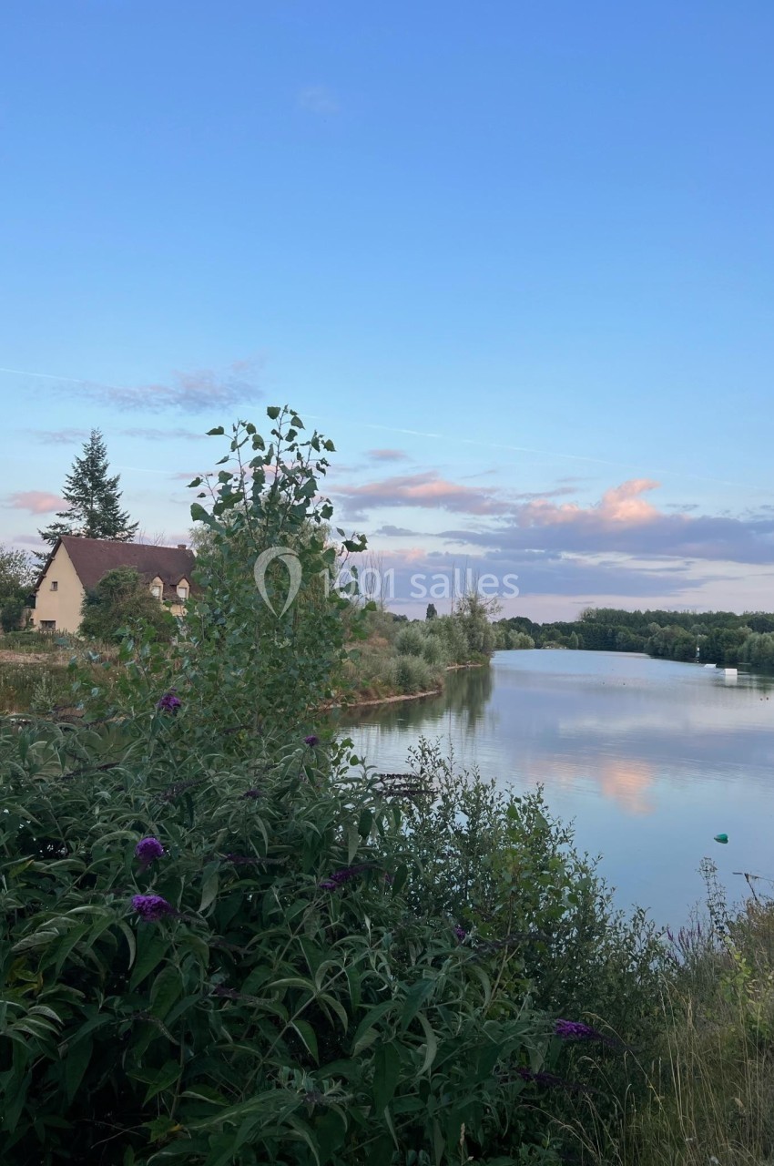 Vue d'une rivière bordée de végétation avec une maison au toit rouge et un ciel partiellement nuageux au coucher du soleil.