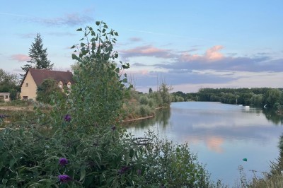 Vue d'une rivière bordée de végétation avec une maison au toit rouge et un ciel partiellement nuageux au coucher du soleil.