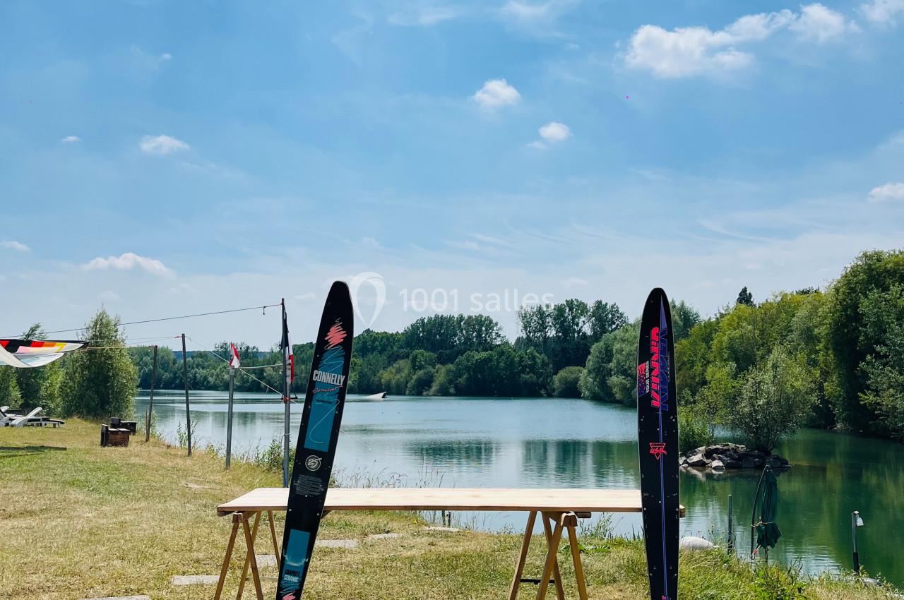 Deux skis nautiques posés contre une table en bois, près d'un lac entouré de verdure sous un ciel dégagé.