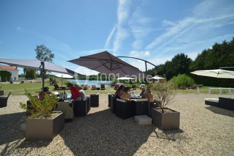Des personnes assises à des tables sous des parasols dans un espace extérieur ensoleillé, près d'un plan d'eau.