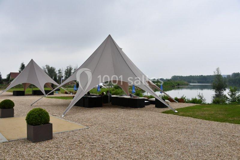 Tentes blanches en étoile installées sur un sol gravillonné près d'un lac entouré de verdure.
