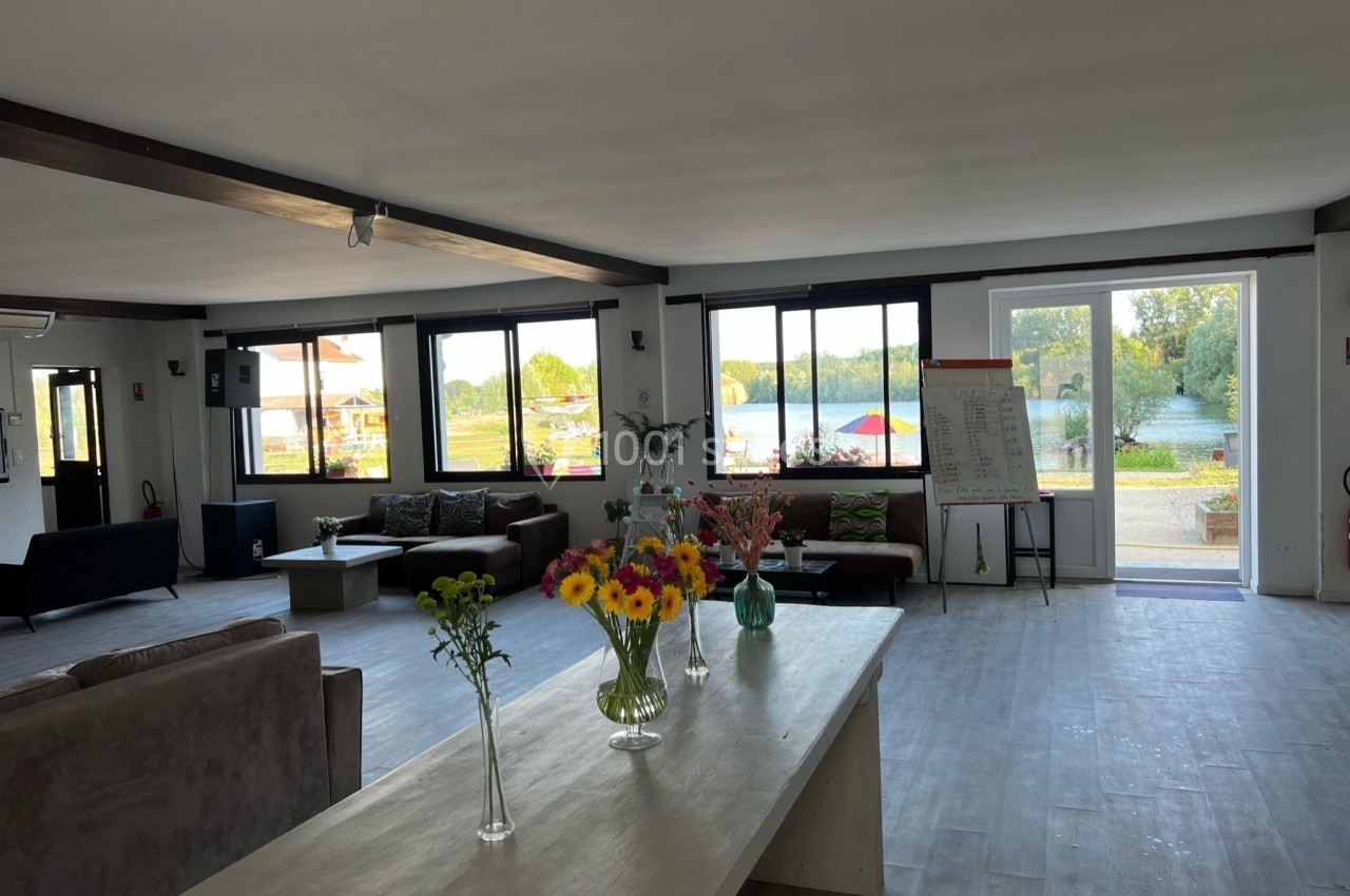 Salle lumineuse avec grandes baies vitrées, mobilier moderne, fleurs sur une table et vue sur un espace extérieur verdoyant.