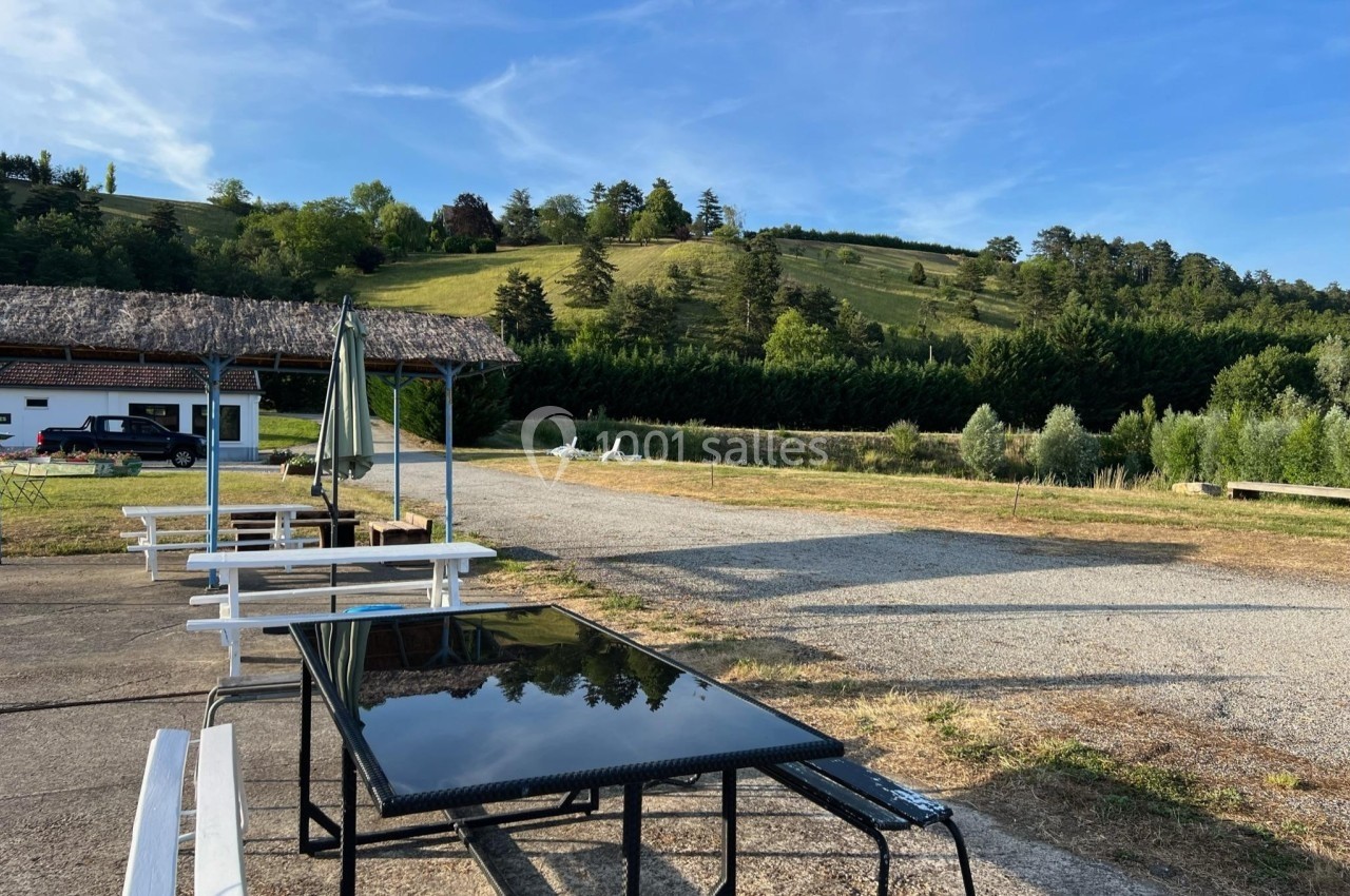 Tables et bancs en plein air près d'un bâtiment, avec une colline boisée en arrière-plan sous un ciel dégagé.