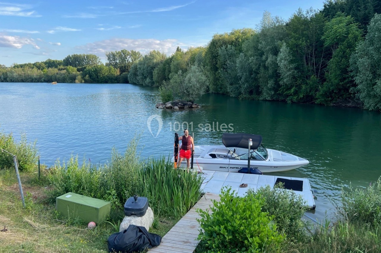 Un homme debout sur un ponton en bois près d'un bateau amarré, entouré de végétation et d'un lac calme.