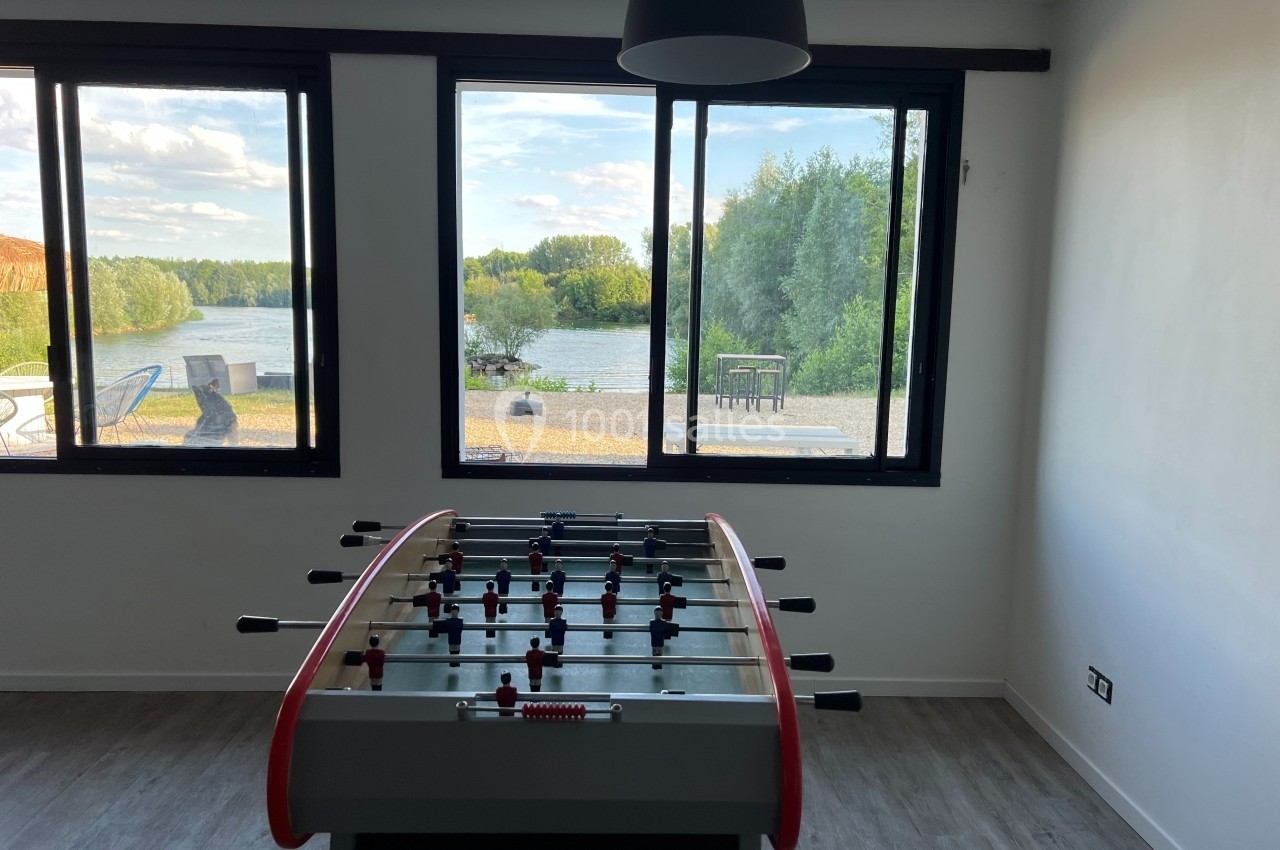 Table de baby-foot dans une pièce lumineuse avec vue sur un lac et des arbres à travers de grandes fenêtres.