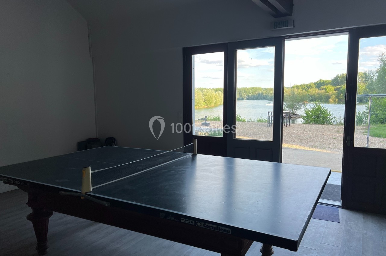 Table de ping-pong dans une pièce lumineuse avec vue sur un lac et des arbres à travers des portes vitrées.