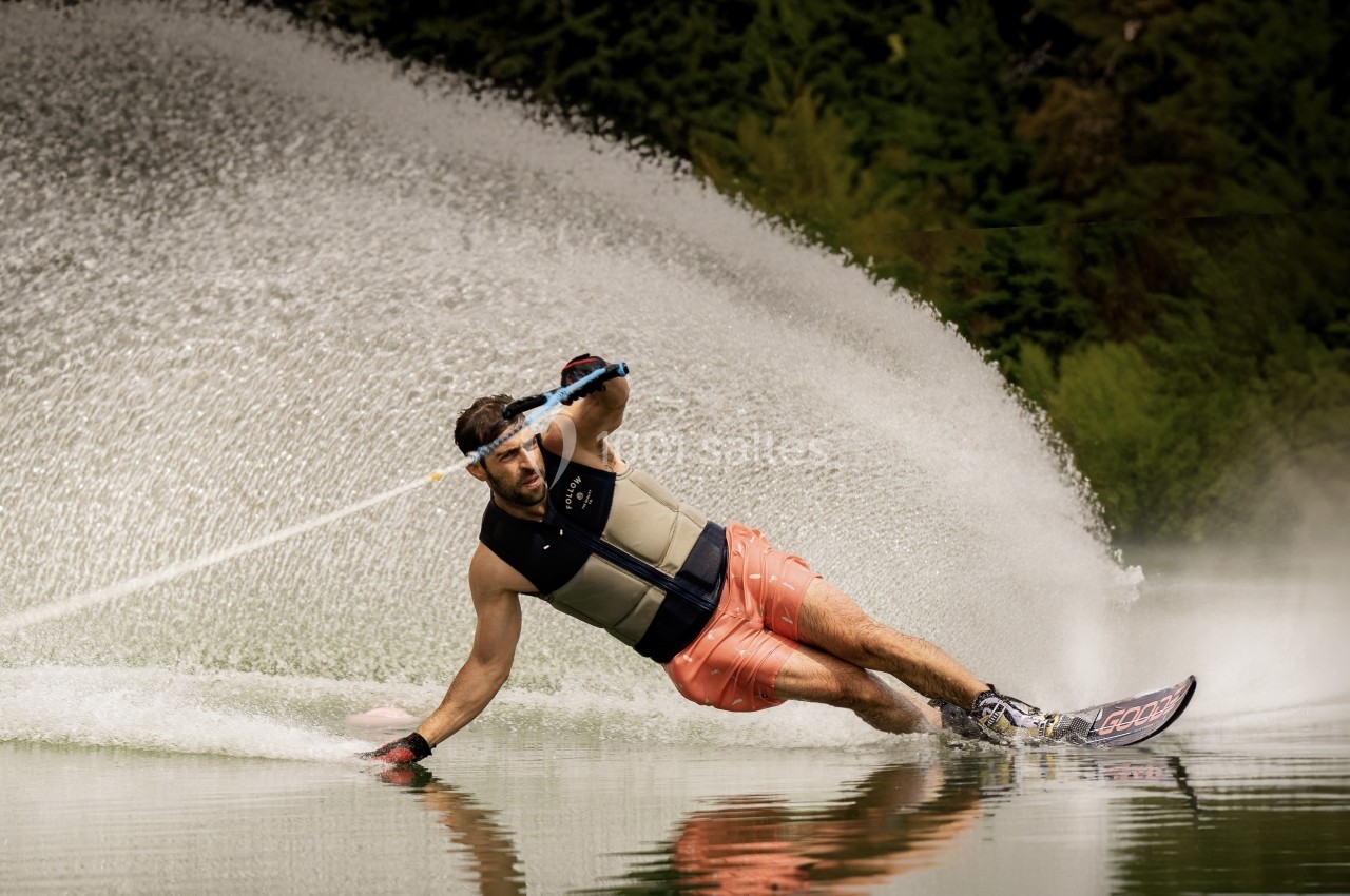 Un homme en gilet de sauvetage pratique le ski nautique, créant une gerbe d'eau sur un lac entouré de végétation.