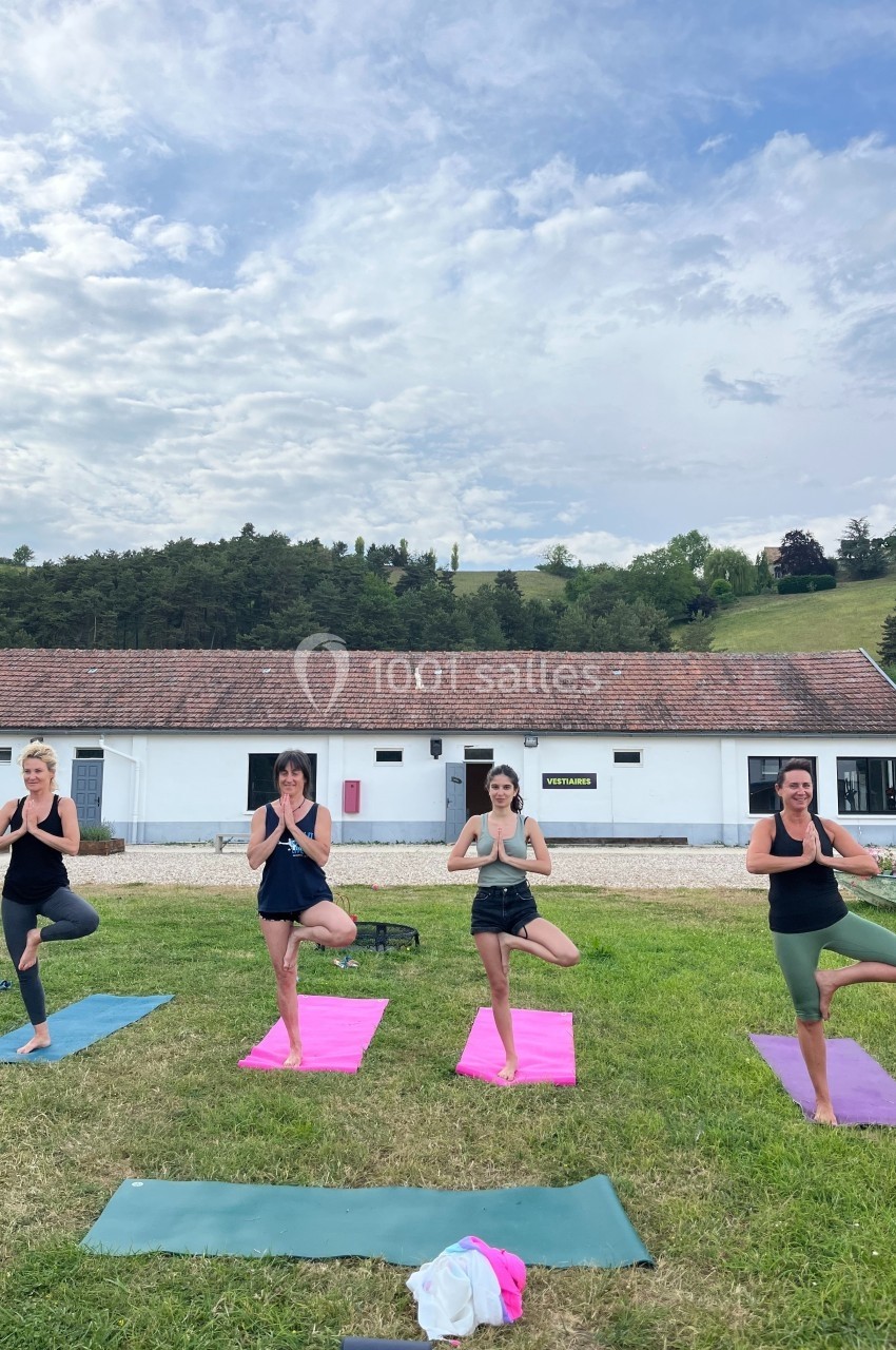 Quatre personnes pratiquent le yoga en posture de l'arbre sur des tapis colorés dans un espace extérieur herbeux.