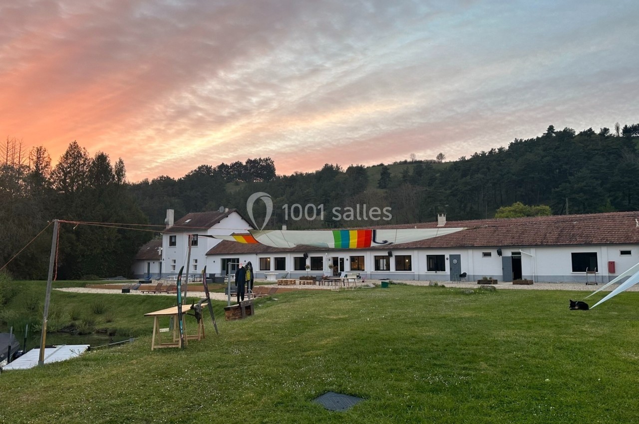 Bâtiment blanc à un étage avec une banderole colorée, entouré de verdure, sous un ciel de coucher de soleil.