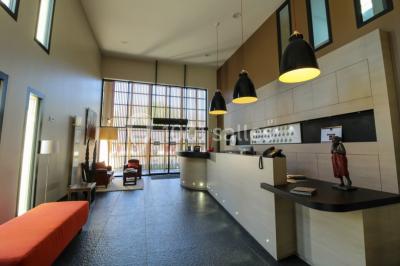 Salon d'hôtel avec canapés beiges, table basse, bureau, chaise noire et grande baie vitrée donnant sur un jardin.