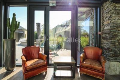 Salon d'hôtel avec canapés beiges, table basse, bureau, chaise noire et grande baie vitrée donnant sur un jardin.