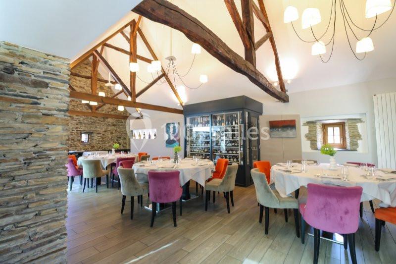 Salle de restaurant lumineuse avec poutres apparentes, murs en pierre, tables dressées et chaises colorées.
