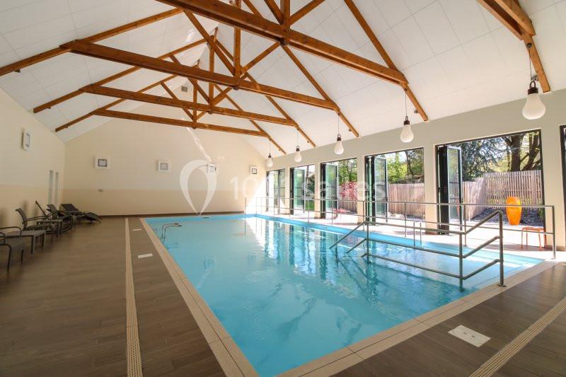 Piscine intérieure lumineuse avec plafond en bois, rampes d'accès et chaises longues sur le côté.