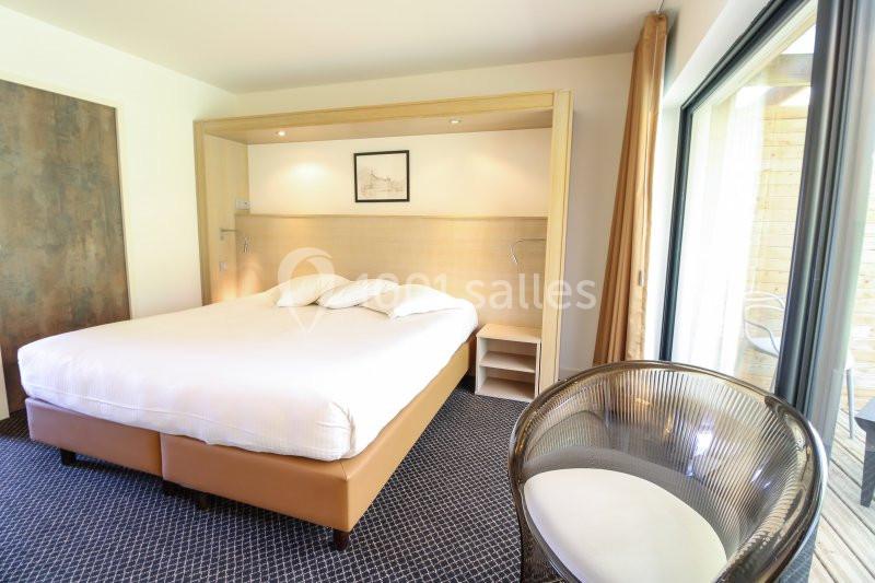 Chambre lumineuse avec un grand lit, un fauteuil en osier, un mur clair et une baie vitrée donnant sur une terrasse.