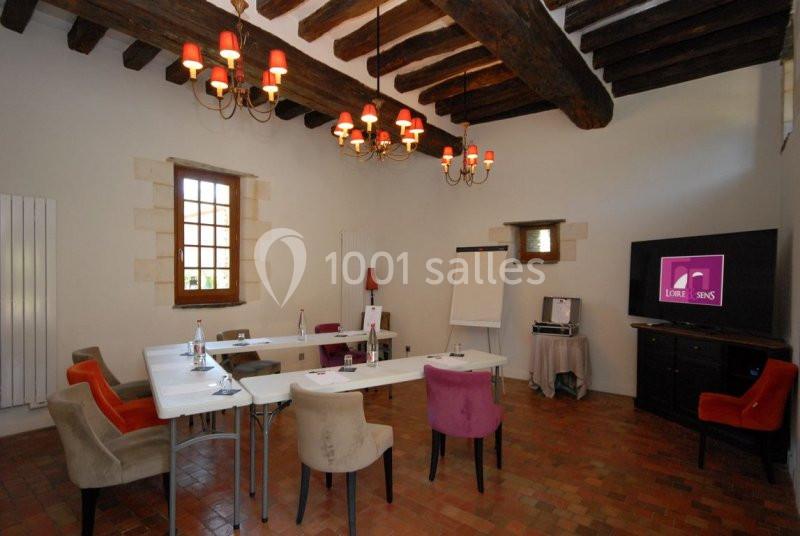 Salle de réunion avec poutres apparentes, tables blanches, chaises colorées, écran, paperboard et éclairage chaleureux.