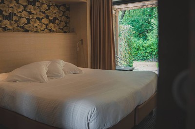 Salon d'hôtel avec canapés beiges, table basse, bureau, chaise noire et grande baie vitrée donnant sur un jardin.