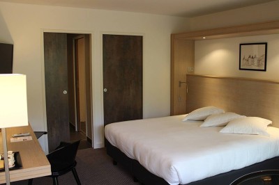 Salon d'hôtel avec canapés beiges, table basse, bureau, chaise noire et grande baie vitrée donnant sur un jardin.