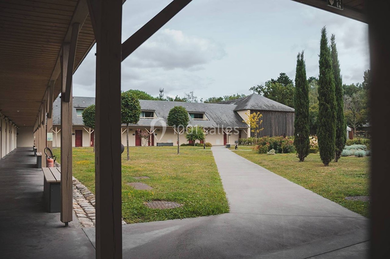 Allée en béton bordée de pelouses et d'arbres, menant à un bâtiment avec toit en ardoise et arcades en bois.