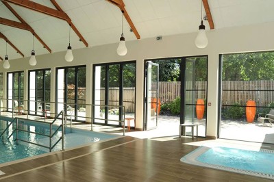 Salon d'hôtel avec canapés beiges, table basse, bureau, chaise noire et grande baie vitrée donnant sur un jardin.