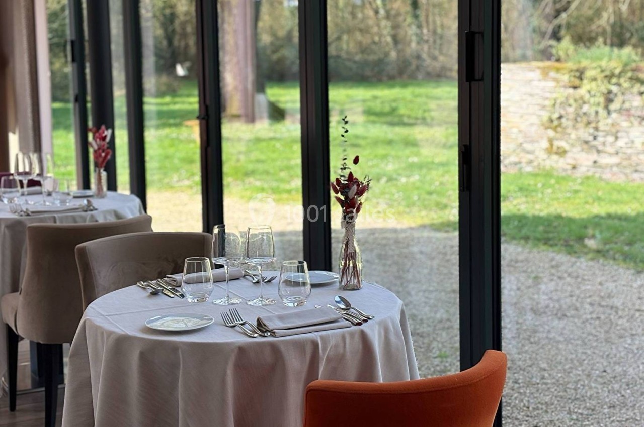 Tables dressées avec nappes blanches et vaisselle élégante, près de grandes baies vitrées donnant sur un jardin verdoyant.
