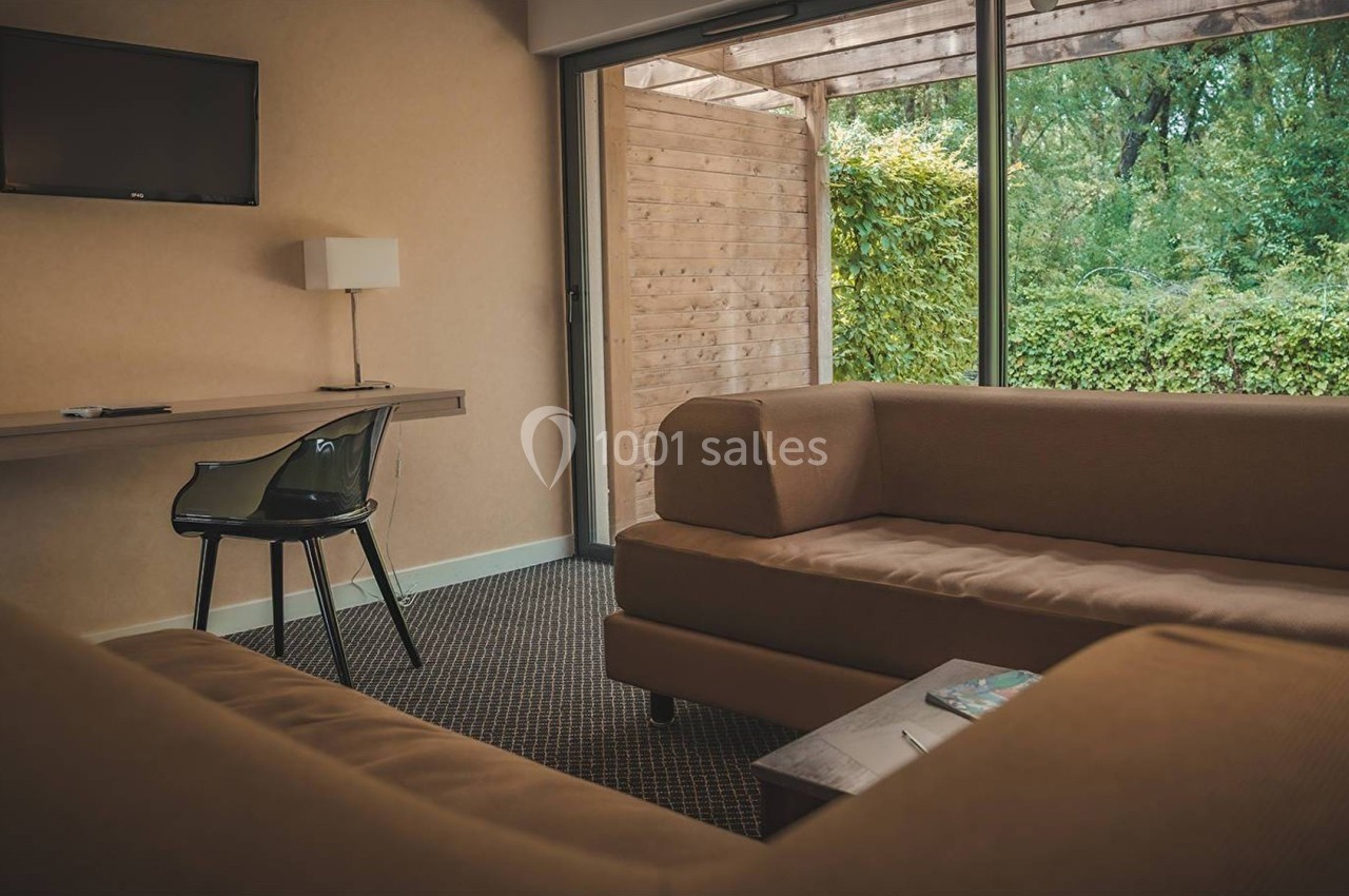Salon d'hôtel avec canapés beiges, table basse, bureau, chaise noire et grande baie vitrée donnant sur un jardin.