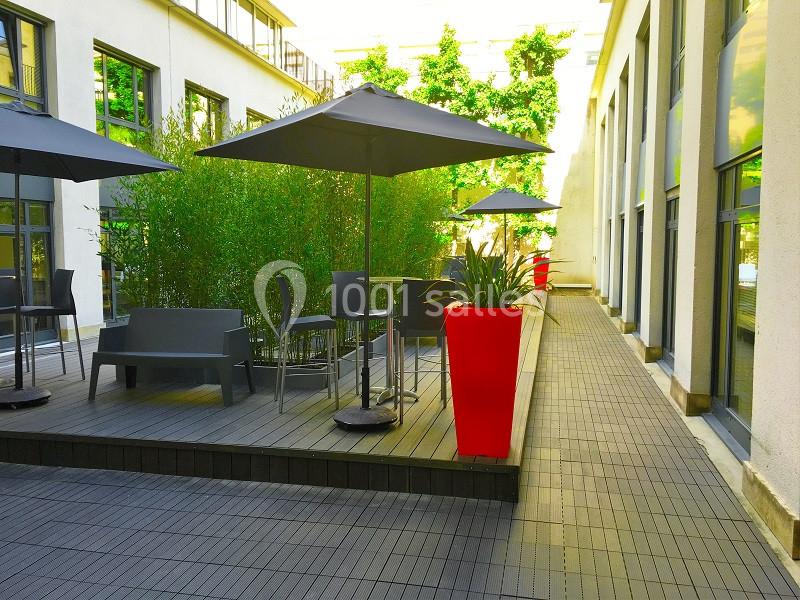 Terrasse extérieure avec tables, chaises, parasols, plantes en pots rouges et bambous, entourée de bâtiments modernes.