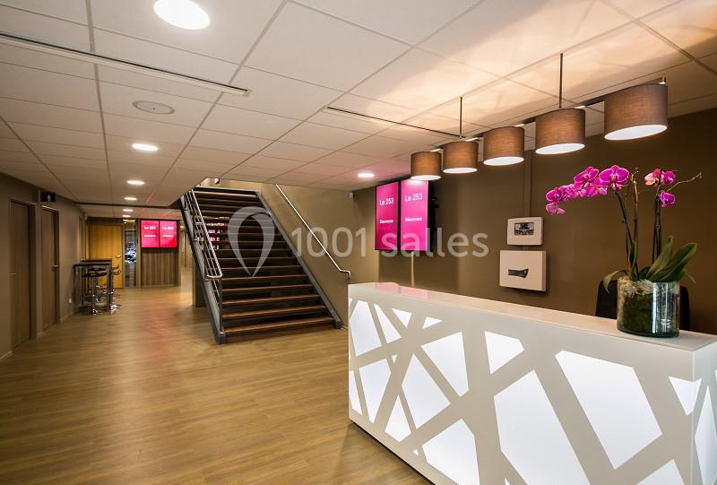 Réception moderne avec comptoir blanc, escalier en bois, écrans rouges et orchidée rose dans un espace éclairé.