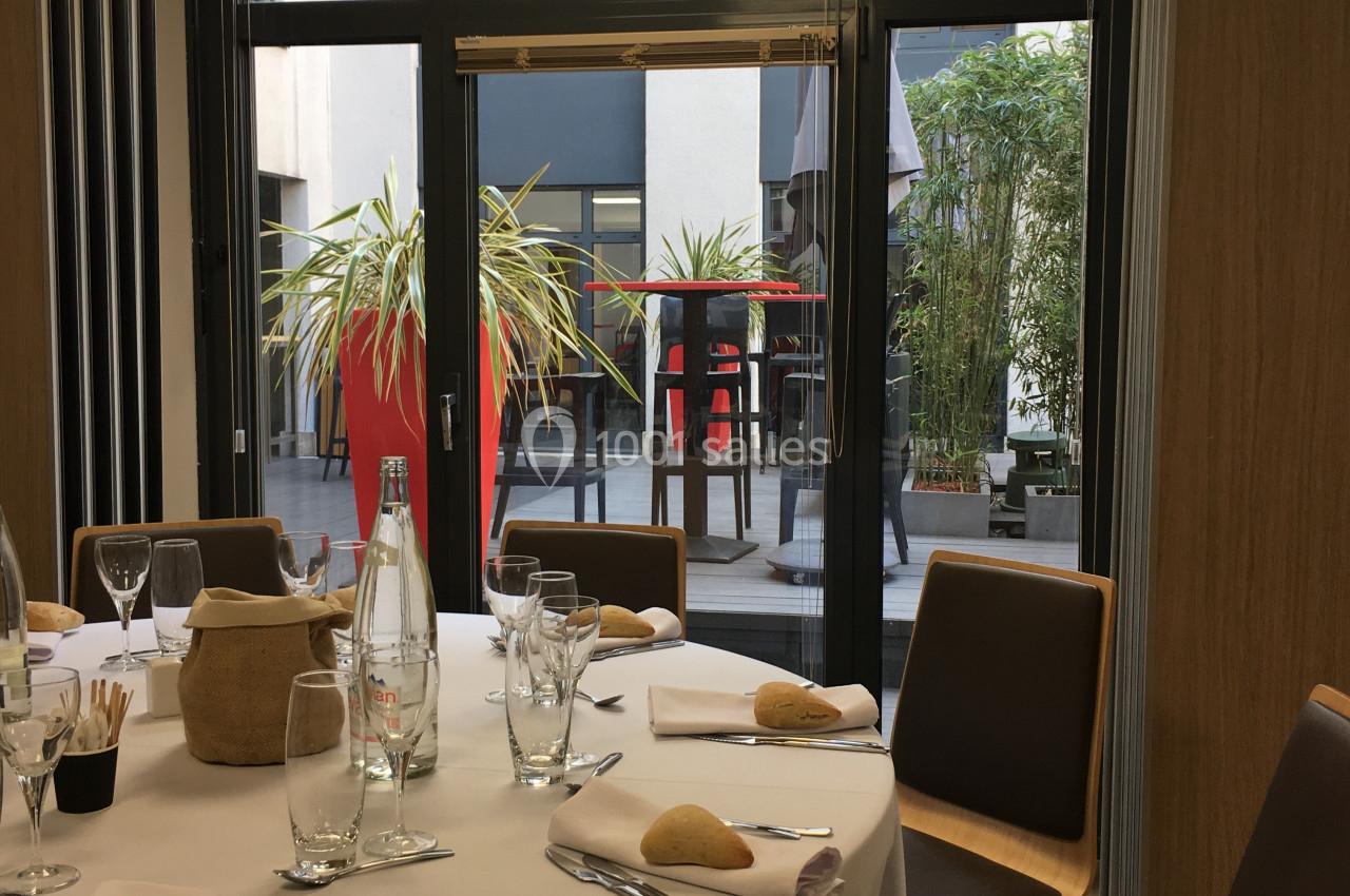 Salle de réunion avec table dressée pour un repas, vue sur une terrasse avec plantes et mobilier extérieur.