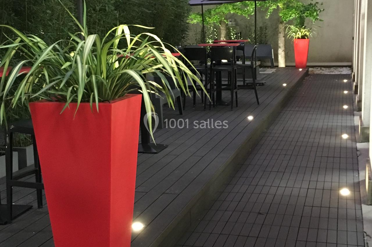Terrasse en bois avec tables, chaises, parasols gris et plantes en pots rouges, éclairée par des lumières au sol.