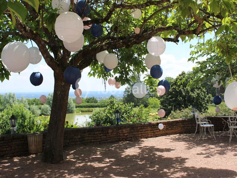 Lanternes suspendues dans un arbre sur une terrasse en gravier, avec vue sur un jardin et un paysage verdoyant.