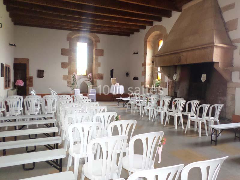 Salle avec des rangées de chaises blanches, une grande cheminée en pierre et des fenêtres laissant entrer la lumière.