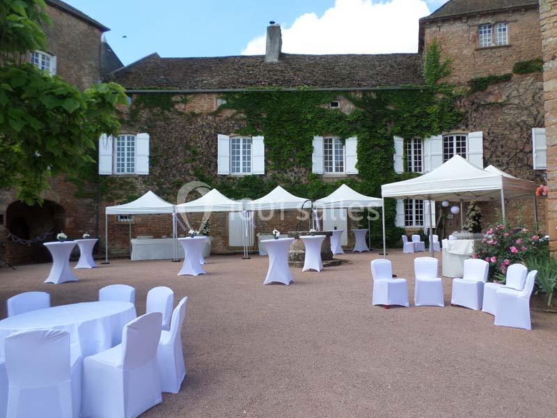 Cour d’un bâtiment en pierre avec des tentes blanches, des tables et des chaises habillées pour un événement extérieur.