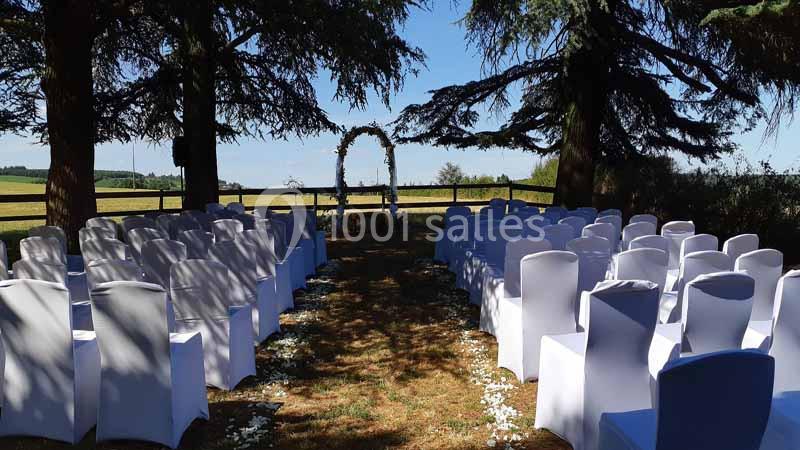 Chaises blanches alignées en extérieur sous des arbres, formant une allée menant à une arche décorée pour une cérémonie.