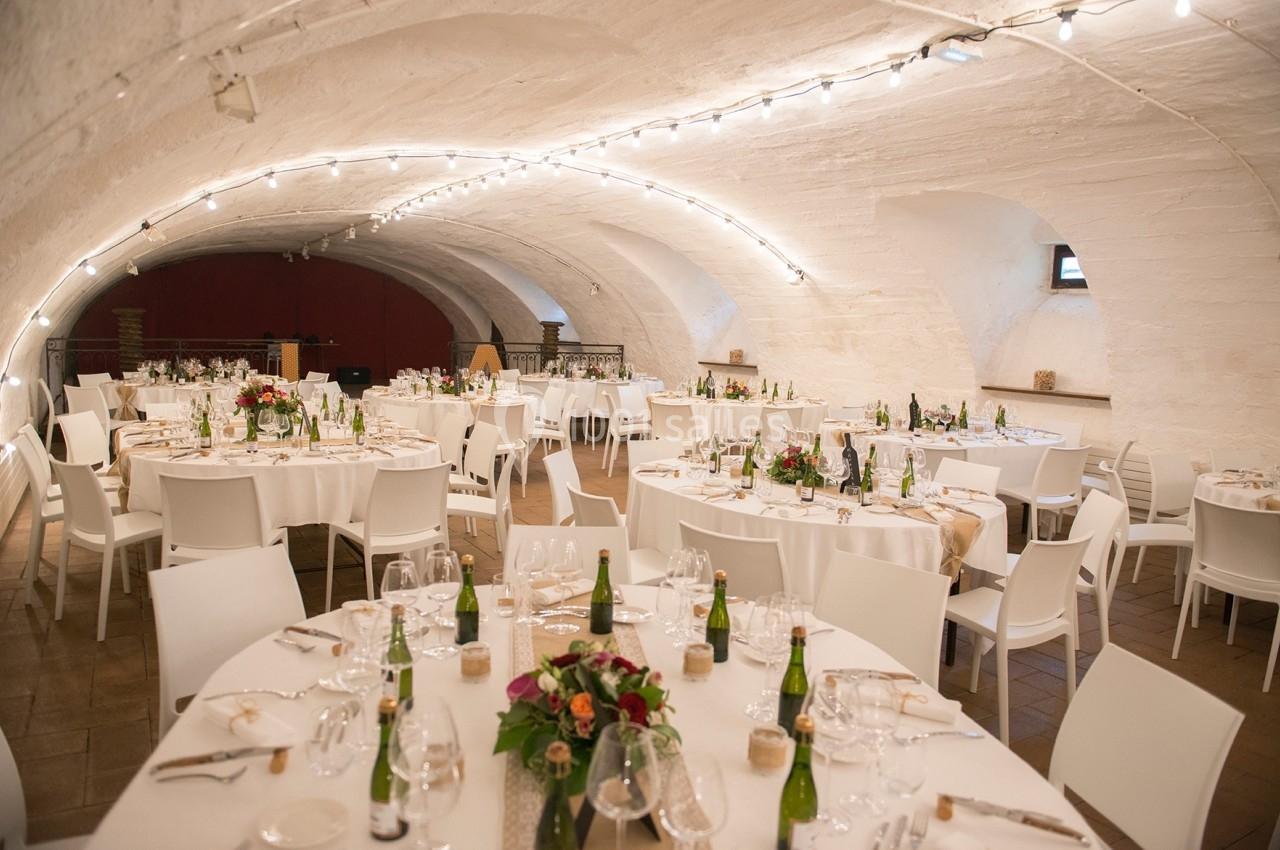 Salle voûtée décorée pour un événement, avec des tables rondes dressées, des fleurs et des bouteilles disposées.
