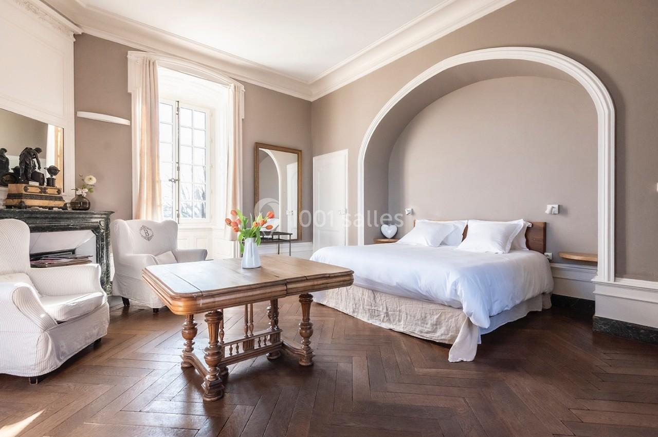 Chambre lumineuse avec lit double, parquet en bois, table basse, fauteuils blancs et vase de fleurs sur la table.