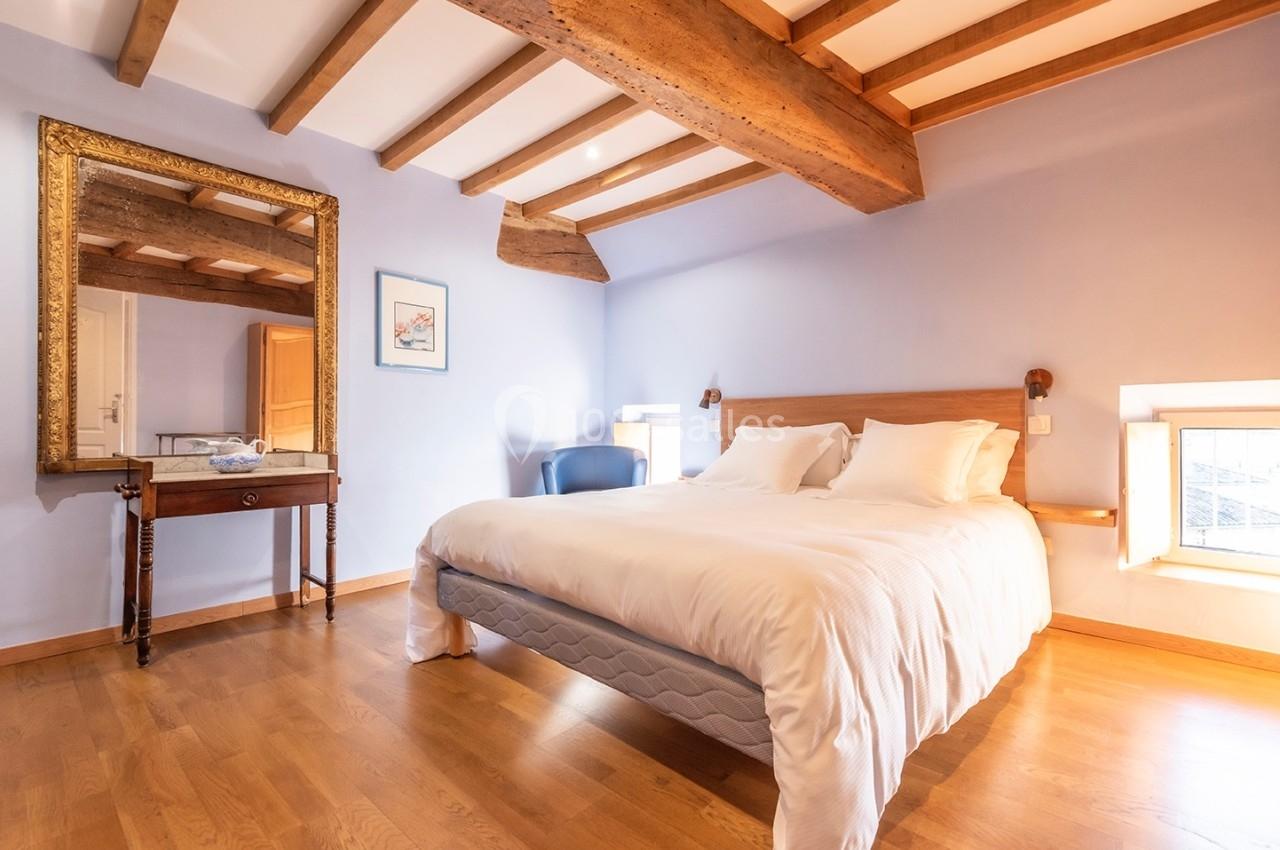 Chambre lumineuse avec lit double, poutres apparentes, parquet en bois, miroir doré et bureau ancien.
