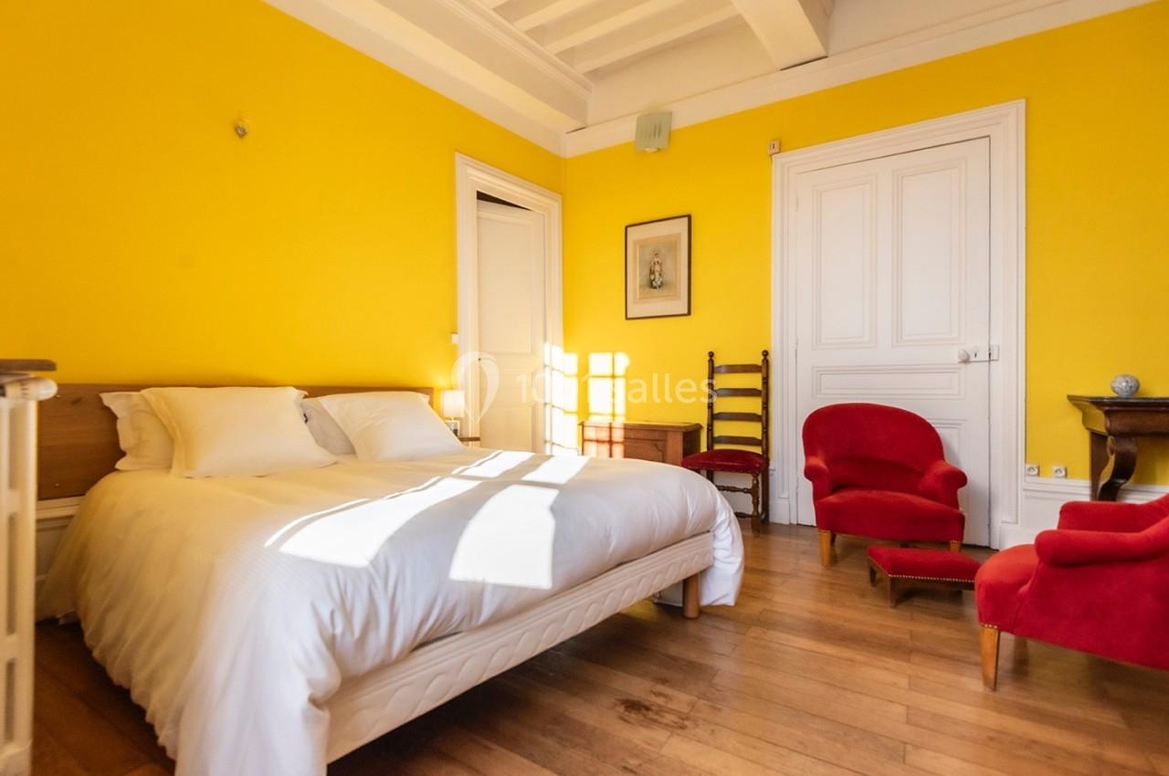 Chambre lumineuse avec murs jaunes, lit double, fauteuils rouges, parquet en bois et mobilier classique.